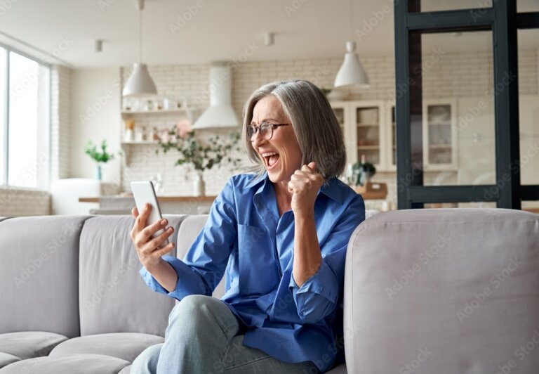 Eine ältere Frau mit grauen Haaren sitzt lächelnd auf dem Sofa und nutzt ein Smartphone.